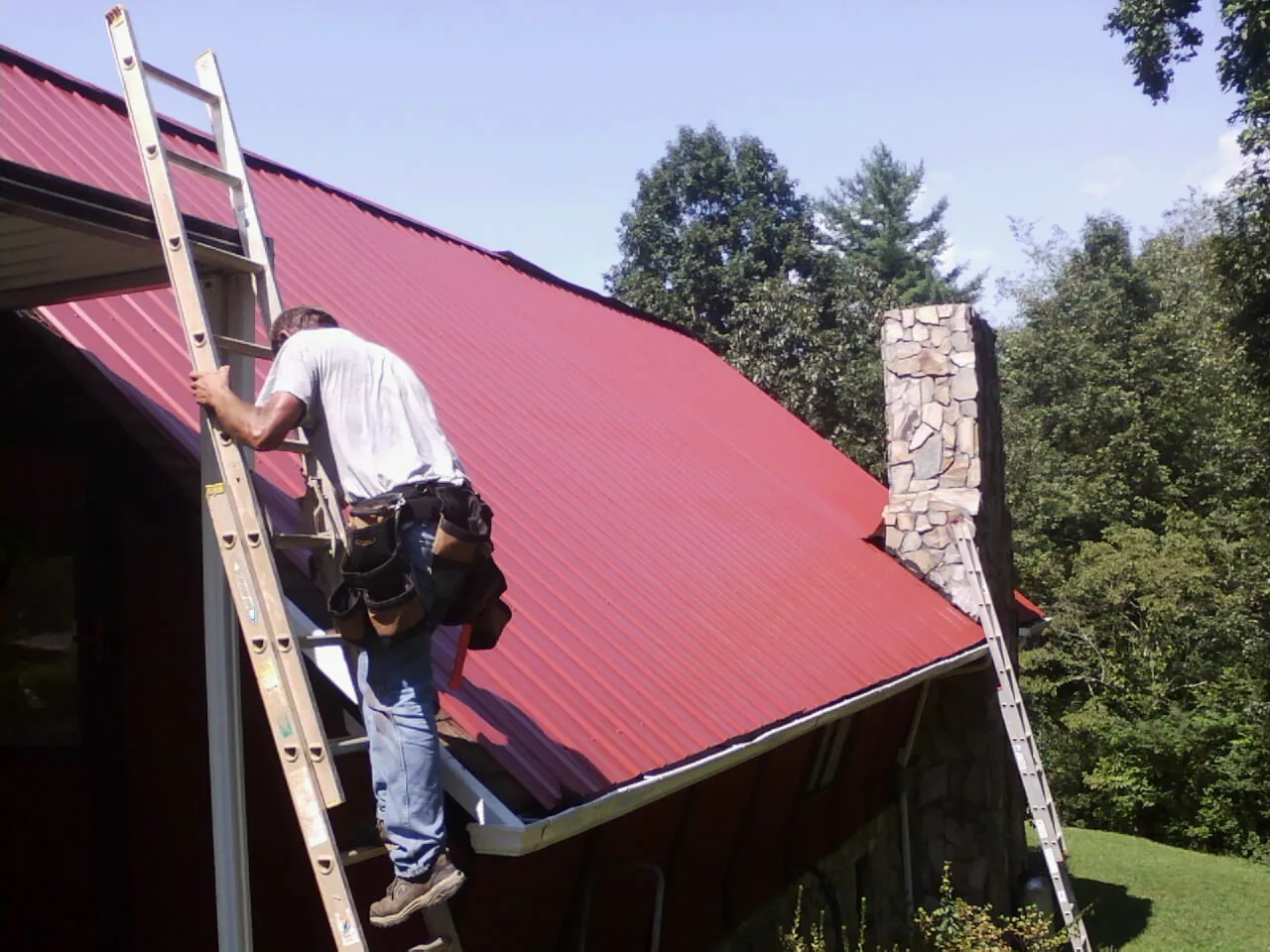 Roofer on a ladder beside a red metal roof and stone chimney — Soffit & Fascia Repair by Roofing Brothers in Waltersburg